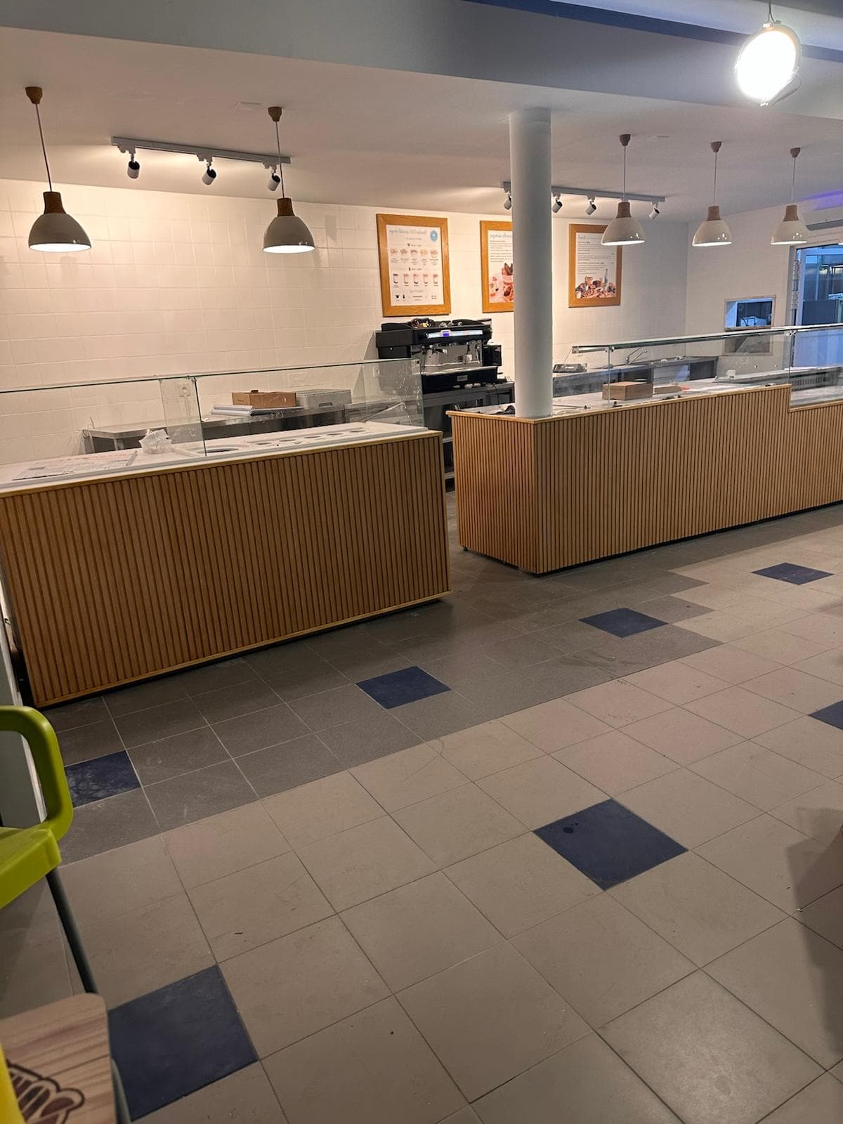 Cafetaria em obra com novo balcão em ripado de madeira já instalado, candeeiros suspensos brancos e cartazes na parede.