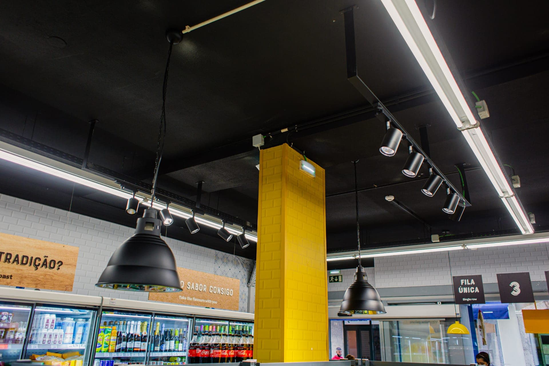 Interior remodelado do supermercado Mini Preço da Baixa-Chiado, com luminárias industriais pretas suspensas e coluna central em mosaico amarelo.