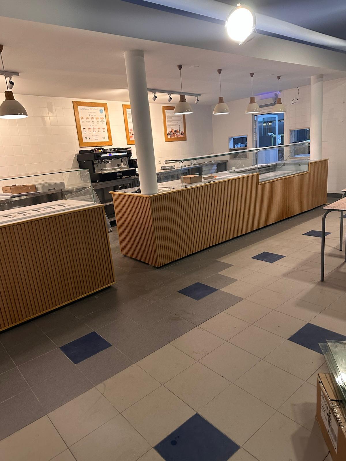 Vista lateral da cafetaria em obra com candeeiros pendentes e quadros de menu na parede.