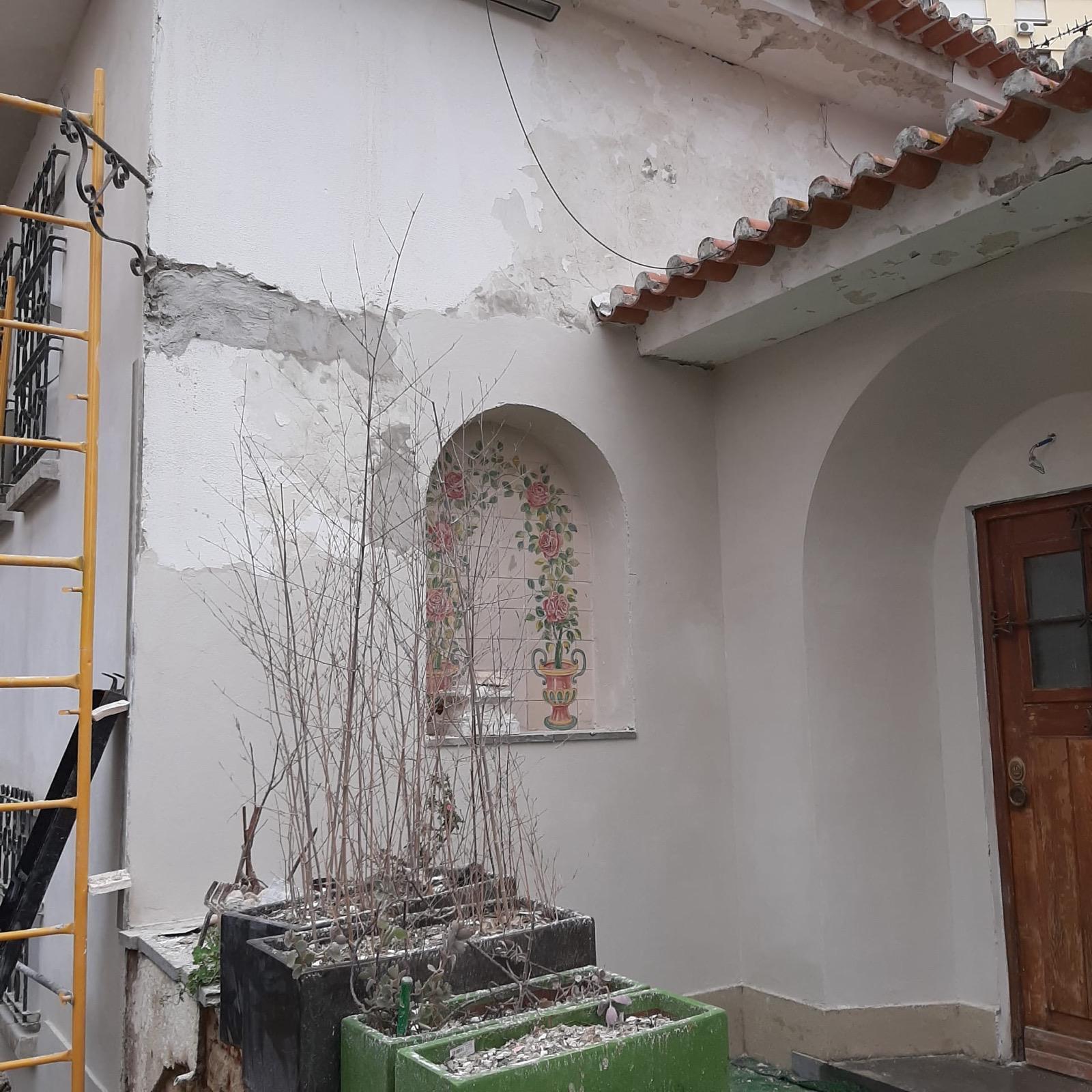 Pátio de entrada com mesa em madeira, janelas restauradas e arbustos do jardim.