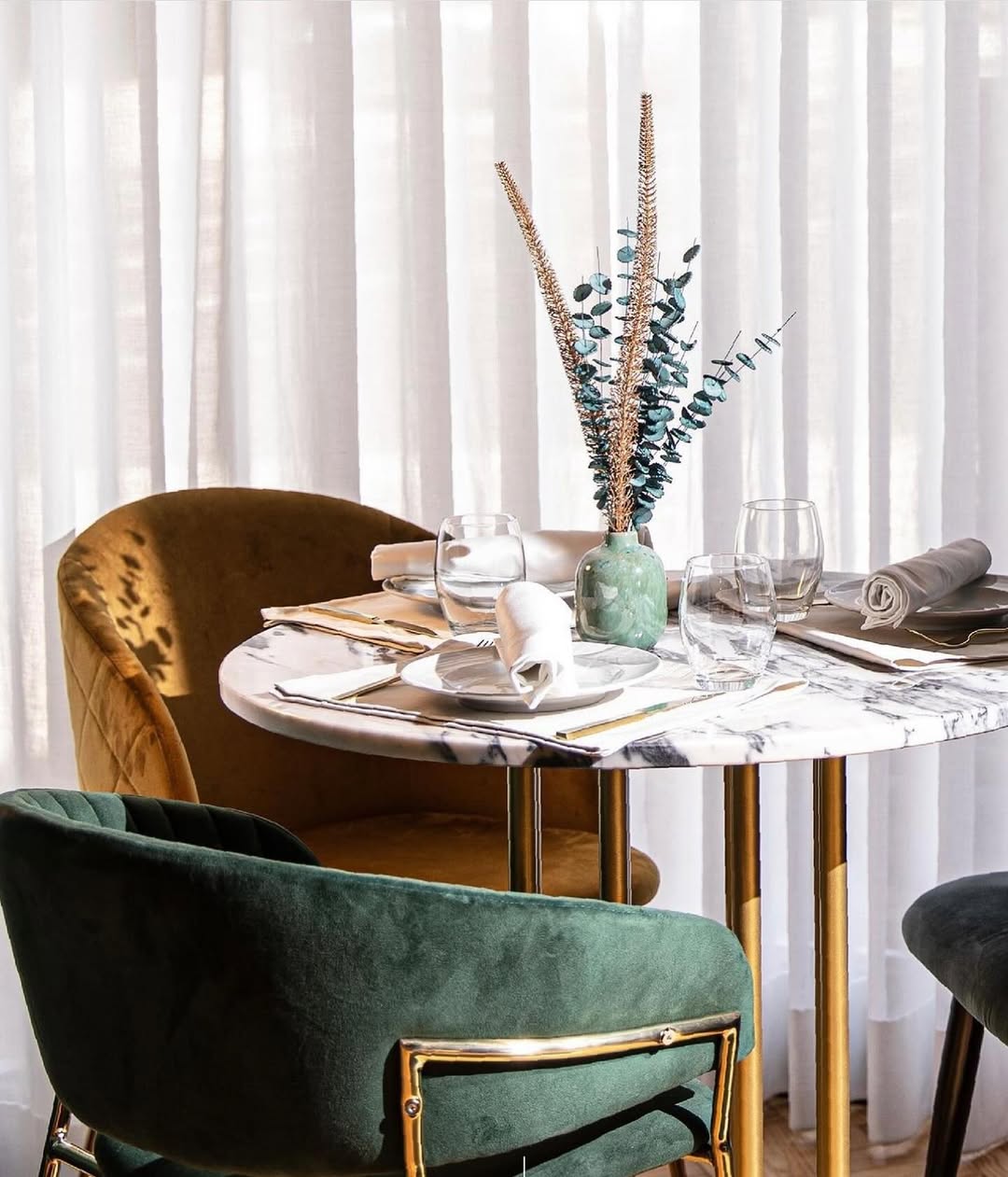 Detalhe de mesa redonda em mármore com cadeiras em veludo verde e amarelo, junto a cortinas brancas.