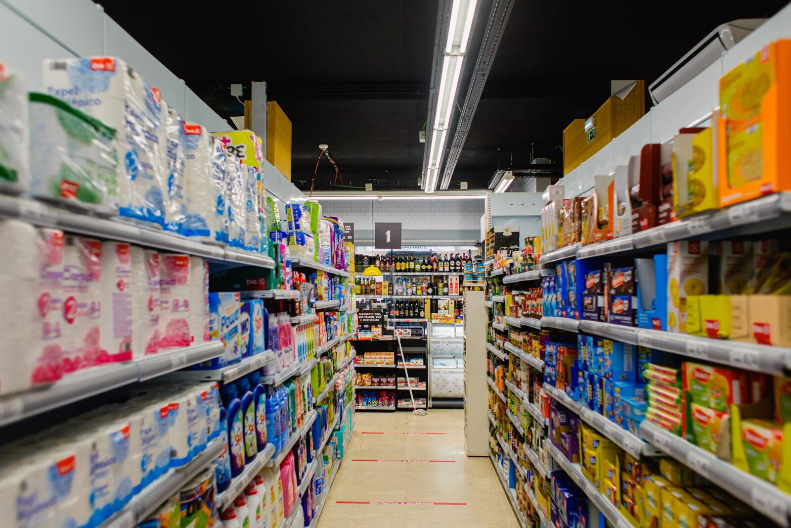 Corredor principal do supermercado com iluminação linear e prateleiras carregadas.