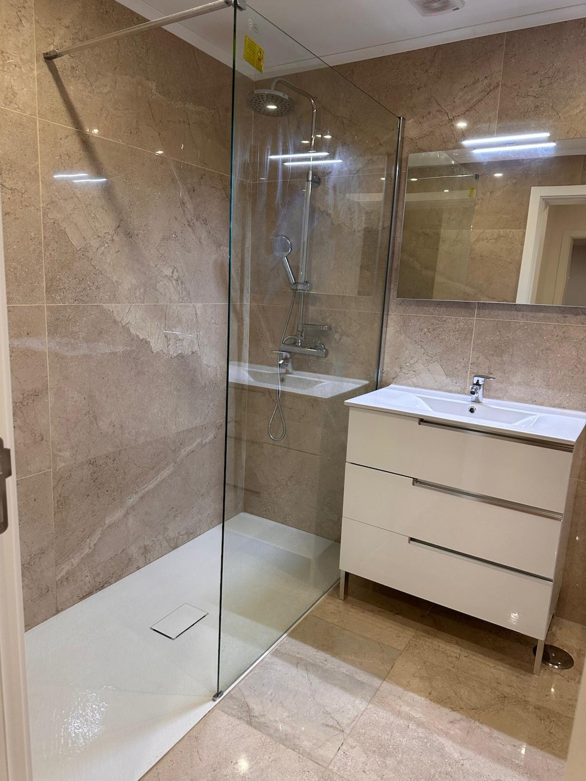Casa de banho nova em mármore travertino, chuveiro com painel de vidro e mobiliário branco.